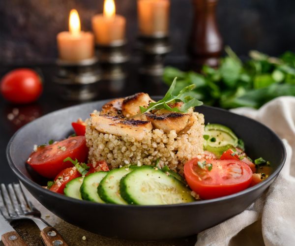 Firefly Quinoa Salad with Grilled Chicken, Tomatoes, and Cucumbers in a black bowl with candles in t